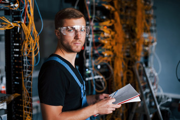 Wall Mural - Young man in uniform with notepad in hands works with internet equipment and wires in server room
