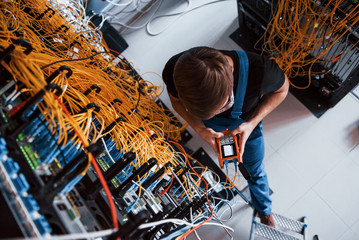 Wall Mural - Top view of young man in uniform with measuring device that works with internet equipment and wires in server room