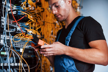 Wall Mural - Young man in uniform with measuring device works with internet equipment and wires in server room