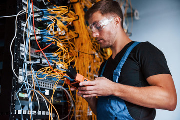 Wall Mural - Young man in uniform with measuring device works with internet equipment and wires in server room