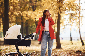 Wall Mural - Talking by the phone. Mother in red coat have a walk with her kid in the pram in the park at autumn time