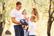 © standret - Mother and dad holds kids at shoulders and in hands. Cheerful young family have a walk in an autumn park together