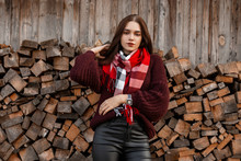 Model Of A Pretty Young Woman In A Vintage Knitted Sweater With A Warm Checkered Trendy Scarf In Leather Pants Posing Near An Old Village Shed. Attractive Pretty Stylish Girl Stands Near Logs Outdoors