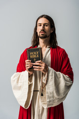 Wall Mural - man holding rosary beads and holy bible and looking at camera isolated on grey