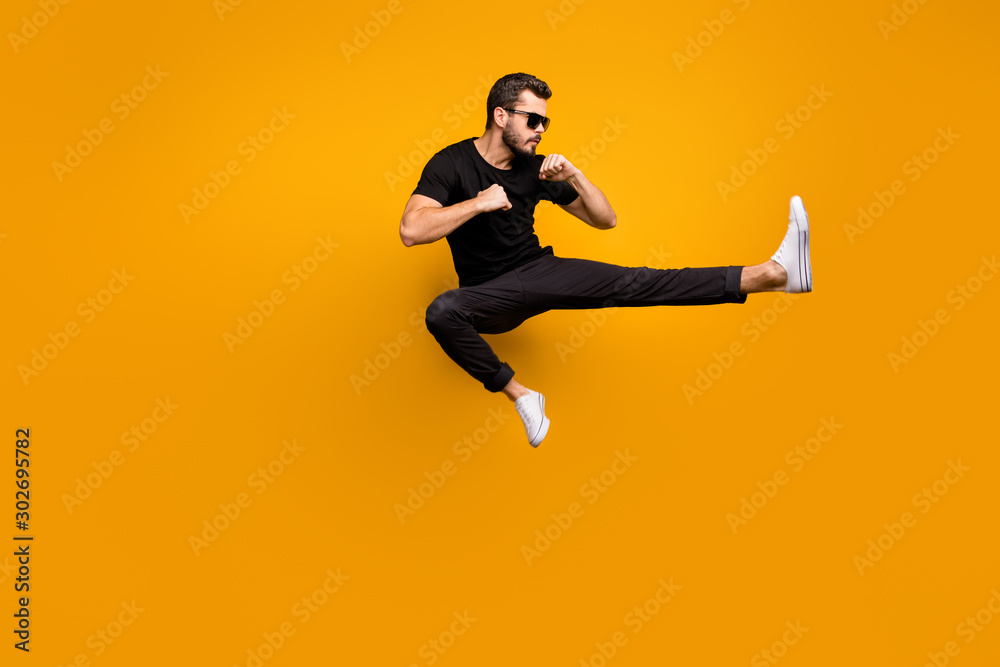 Full length photo of handsome guy jumping high practicing self defense kicking confident facial expression wear sun specs black t-shirt pants isolated yellow color background - obrazy, fototapety, plakaty 