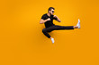Full length photo of handsome guy jumping high practicing self defense kicking confident facial expression wear sun specs black t-shirt pants isolated yellow color background