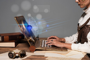 Wall Mural - cropped view of woman typing on steampunk laptop at table with glowing illustration isolated on grey