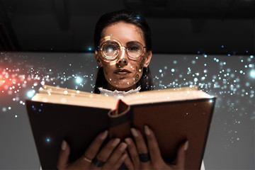 Wall Mural - low angle view of steampunk woman in glasses reading book with glowing illustration above