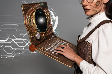 cropped view of steampunk woman using vintage laptop with glowing digital illustration isolated on grey