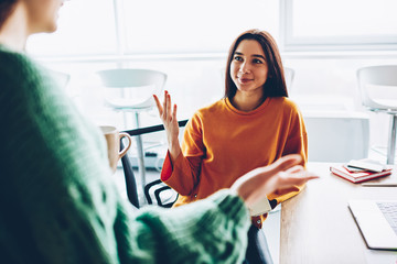  Female designer with brunette hair explaining new creative idea to colleague during break sitting at desktop in university.Attractive young woman dressed in casual wear talking during free time