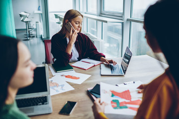 Canvas Print - Positive blonde woman having telephone conversation with operator while using laptop computer in office interior, women crew having productive working process and cooperating using modern technology