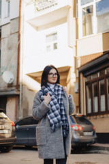 Wall Mural - Attractive positive young girl wearing glasses in a coat on the background of buildings on cars