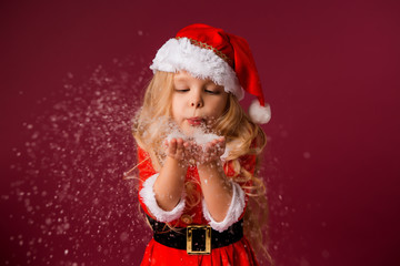 little blonde girl in a Santa suit blows snow off her hands. red background isolate. space for text