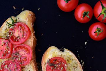 two slices of Italian white bread with green olive oil and sliced ​​tomato with oregano next to fresh red tomatoes against dark background