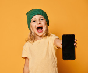 Smiling screaming little girl kid in green modern winter hat showing blank screen of new popular mobile phone