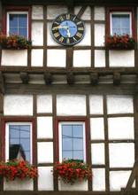 Vertical Shot Of A Building With A Vintage Clock On Top And Red Flowers On The Window Sills