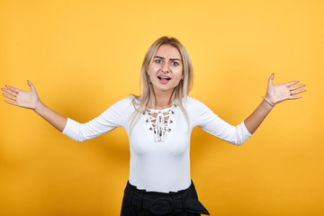 Angry cheerful young woman in casual shirt, looking camera, spreading hands isolated on orange background in studio. People sincere emotions, lifestyle concept.