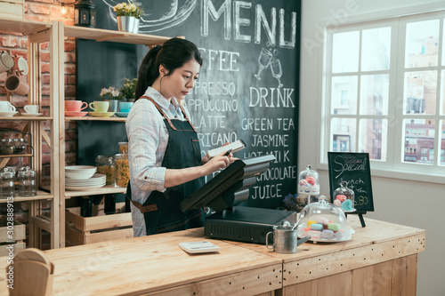 Young asian japanese lady waitress using digital device for payment in coffee shop counter. beautiful woman staff enter customer order from note into point of sale terminal machine in cafe store.