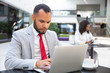 © Mangostar - Focused serious professional using laptop while drinking coffee in office lobby. Young African American woman using tablet in background. Wireless technology concept