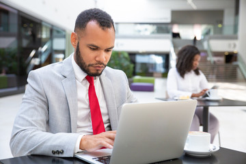 Wall Mural - Focused serious professional using laptop while drinking coffee in office lobby. Young African American woman using tablet in background. Wireless technology concept