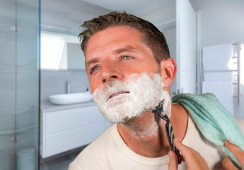 domestic lifestyle morning portrait of attractive and happy man shaving at modern apartment  bathroom his face covered with foam looking on the mirror in daily routine