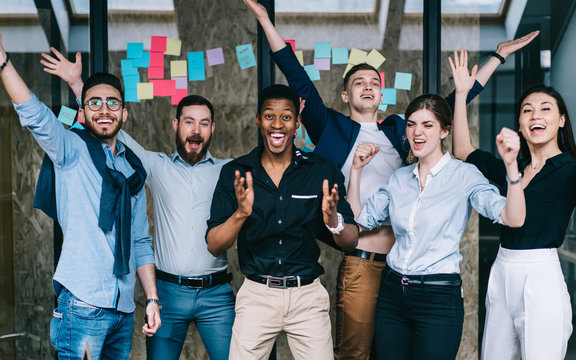 Young crew of happy excited male and female business partners celebrating completed startup project while looking at camera and laughing, overjoyed colleagues feeling amazed of done good work