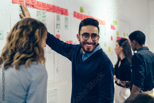 Fotomural Positive young man laughing while collaborating with colleagues on creating presentation using colorful stickers for productive work in office