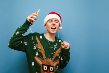 Cheerful Christmas guy dancing in headphones and with smartphone in his hand against a blue background, listening to music with his eyes closed, wearing a santa hat and a warm sweater. Isolated.