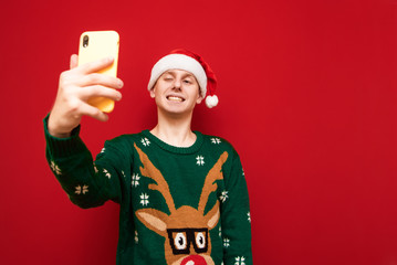 Wall Mural - Smiling guy in santa hat and christmas sweater takes selfie on smartphone, smiles and looks into camera. isolated on red background. Happy young man making christmas selfie on smartphone.