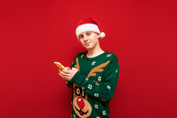 Positive guy in christmas clothes uses smartphone on red background, looks into camera and smiles, wears hat of santa claus and warm green sweater. Isolated. Copy space