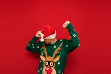 Cheerful young man in santa hat and green sweater dancing on red background. Cheerful guy having fun at Christmas party. Isolated. Copy space