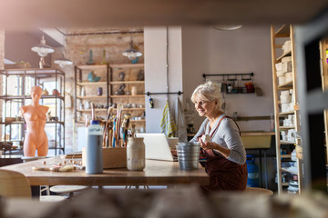 Wall Mural - Mature woman pottery artist using laptop in art studio 