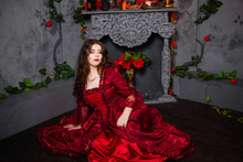 A Beautiful Girl In A Magnificent Red Dress Of The Rococo Era Sits On The Floor Near A Fireplace And Flowers.