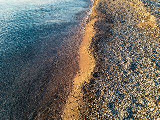 Wall Mural - The coastline is covered with pebbles and sand with a sea wave of clear water running onto it, illuminated by yellow sunlight at sunset on an autumn evening.