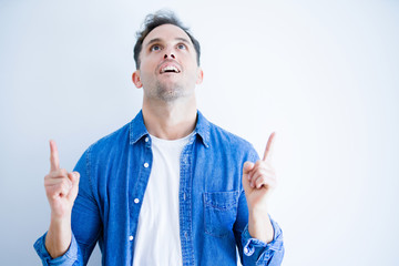 Young handsome man wearing denim shirt standing over isolated white background amazed and surprised looking up and pointing with fingers and raised arms.