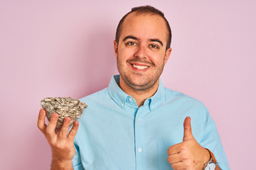 Sticker - Young man holding bowl with sunflowers seeds standing over isolated pink background happy with big smile doing ok sign, thumb up with fingers, excellent sign