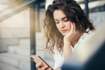 Serious thoughtful young woman sit on steps upset. Look on phone in hands. Sun shines in picture. Sitting alone outside. Brunette posing on camera.