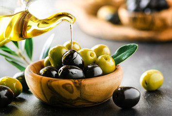 Wall Mural - Pouring fresh virgin olive oil on green and black ripe olives in bowl, on dark stone table or black background.