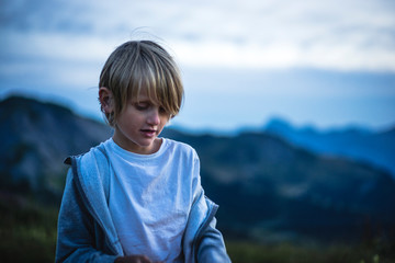 At dusk, portrait of a 7 years old kid looking down, mountain landscape in the background