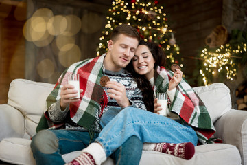 Wall Mural - Young couple beautiful woman and handsome man wearing warm sweaters drinking milk with cookies in room decorated for celebrating the new year christmas festive mood