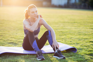 Wall Mural - Young sportive woman in sports clothes sitting on training mat before doing exercises in field at sunrise.