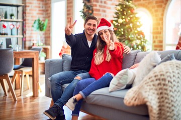 Canvas Print - Young couple wearing santa claus hat sitting on the sofa around christmas tree at home smiling with happy face winking at the camera doing victory sign. Number two.