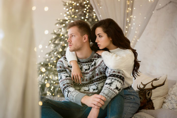 Wall Mural - lovely young couple beautiful woman and handsome man both wearing cozy sweaters hugging on the sofa decorated for celebrating the new year christmas festive mood