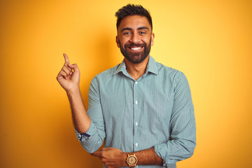 Wall Mural - Young indian man wearing green striped shirt standing over isolated yellow background with a big smile on face, pointing with hand and finger to the side looking at the camera.