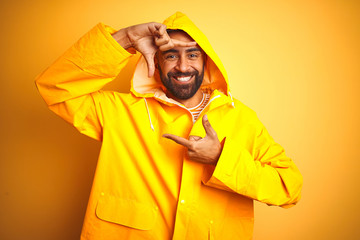 Wall Mural - Young indian man wearing raincoat with hood standing over isolated yellow background smiling making frame with hands and fingers with happy face. Creativity and photography concept.