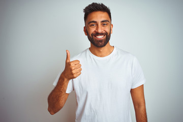 Wall Mural - Young indian man wearing t-shirt standing over isolated white background doing happy thumbs up gesture with hand. Approving expression looking at the camera showing success.