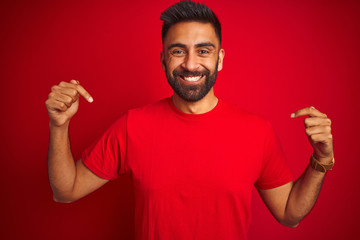 Sticker - Young handsome indian man wearing t-shirt over isolated red background looking confident with smile on face, pointing oneself with fingers proud and happy.