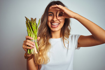 Wall Mural - Young beautiful woman eating asparagus over grey isolated background with happy face smiling doing ok sign with hand on eye looking through fingers