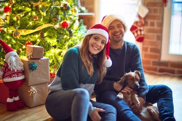 Wall Mural - Young beautiful couple smiling happy and confident.. Sitting on the floor wearing santa claus hat hugging dog around christmas tree at home