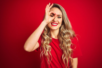 Canvas Print - Young beautiful woman wearing basic t-shirt standing over red isolated background doing ok gesture with hand smiling, eye looking through fingers with happy face.